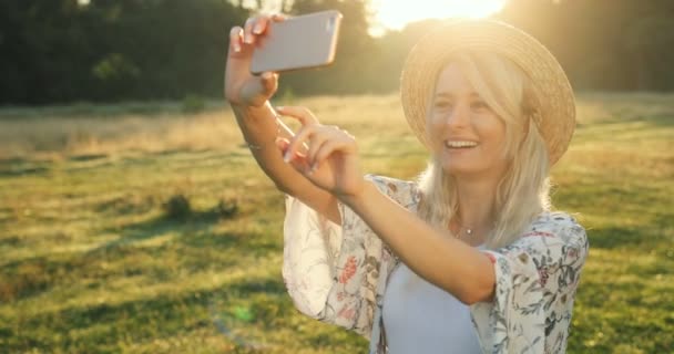 Όμορφη νεαρή ξανθιά γυναίκα στέκεται στο γραφικό χορτοτάπητα και λήψη selfie φωτογραφίες σχετικά με την κάμερα του smartphone. — Αρχείο Βίντεο