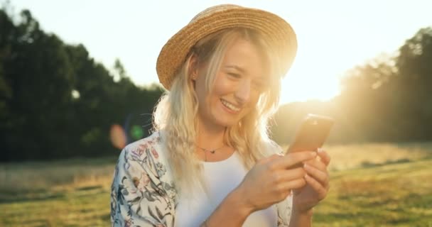 Närbild av den attraktiva unga kaukasiska kvinnan i hatten tejpning medan textning på smartphone-enheten på en solnedgång. Porträtt skott. — Stockvideo