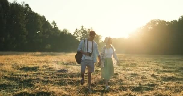 Unga kaukasiska glad flickvän och pojkvän gå hand i hand med en kamera i fältet på sommaren solnedgången. — Stockvideo