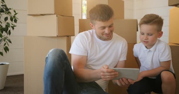 Retrato de fathet e hijo en la tableta, mostrando un juego interesante a su pequeño hijo mientras el hombre le presta atención en el nuevo hogar. Hipoteca, mudanza y concepto de bienes raíces . — Vídeos de Stock