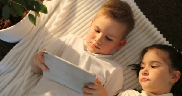 Вид сверху на мальчика и девочку, пользующихся планшетом, лежа на полу. Маленькие дети смотрят на сенсорную панель дома — стоковое видео