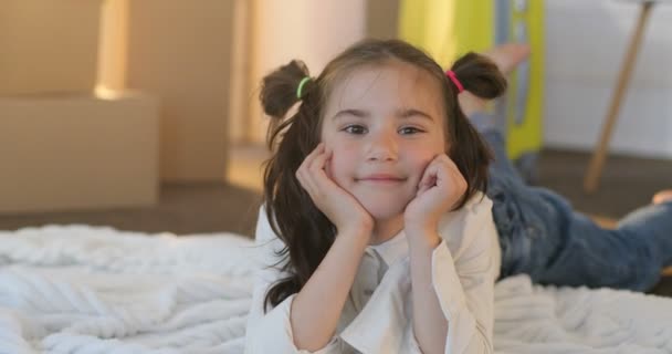 Portrait de mignonne petite fille aux cheveux longs souriant brillamment, tout en étant couché sur le sol à la nouvelle maison, fond de boîtes en carton. Gros plan portrait de mignonne petite fille couchée avec le visage sur les mains dans le parc . — Video