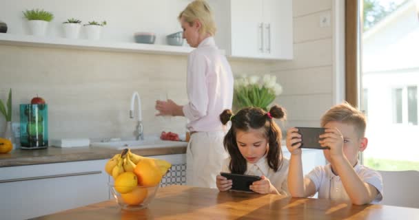 Madre e hijos en la cocina. Pequeña linda chica y niño está utilizando el teléfono inteligente, mientras que la madre está haciendo jugo de la bebida. Concepto familiar — Vídeo de stock