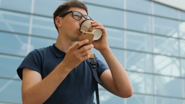 Nahaufnahme Porträt eines jungen Mannes mit Brille, der sein Smartphone benutzt und vor dem Hintergrund des Flughafens Kaffee trinkt. Seitenansicht. — Stockvideo