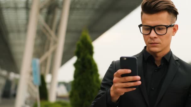 Retrato de jovem homem de negócios nos óculos na elegante camisa preta usando telefone inteligente no fundo do aeroporto . — Vídeo de Stock