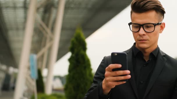 Portret van de jonge zakenman in de glazen in de stijlvolle zwarte shirt met behulp van slimme telefoon op de achtergrond van de luchthaven. — Stockvideo