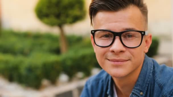 Retrato de jovem feliz homem com óculos olhando para a câmera e sorrindo no fundo da cidade . — Vídeo de Stock