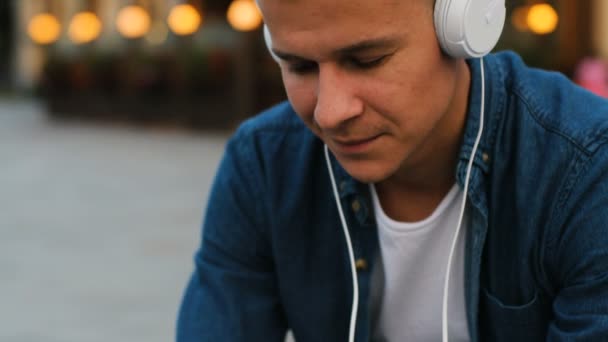 Retrato de un joven en los auriculares escuchando la música mientras se relaja en el banco de la ciudad . — Vídeos de Stock