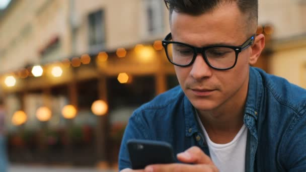 Крупный план привлекательного мужчины в стильных очках, использующего мобильный телефон для общения с друзьями на улице на фоне буке. Портретный снимок . — стоковое видео