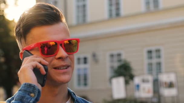 Retrato de un joven atractivo en las gafas de sol hablando en el teléfono inteligente en el fondo de la ciudad . — Vídeos de Stock