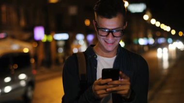 Çekici bir adam akıllı telefon açık renkli üzerinde akşam anda sokakta kullanarak şık bardaklarda görüntüsünü kapatmak.