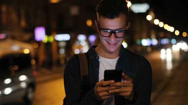 特写镜头的魅力男子在时尚的眼镜使用智能手机在街道上的灯光背景的晚上时间. — 图库视频影像
