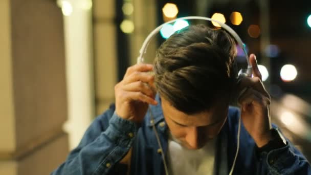 Retrato de un joven con auriculares mientras camina por la calle de la ciudad por la noche y comienza a escuchar música . — Vídeos de Stock