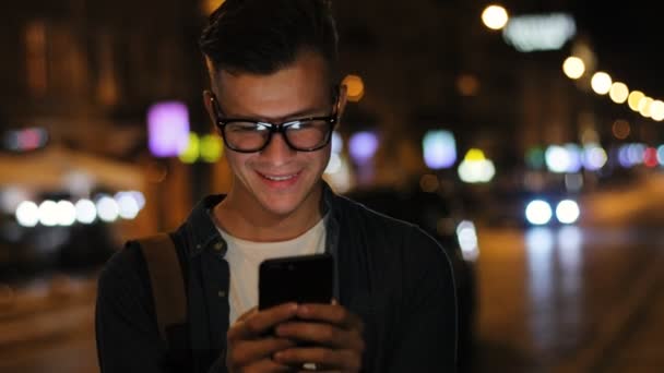Emotionaler Mann mit stylischer Brille am Abend auf der Straße mit Smartphone im Hintergrund. — Stockvideo