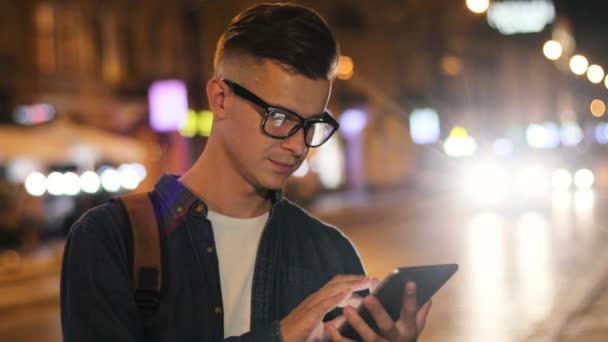 View from the side of emotional man in the stylish glasses using smarphone in the street at the evening time on road background . — стоковое видео