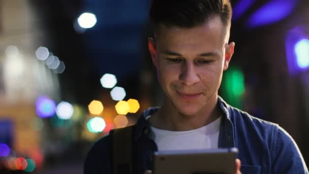 Giovane uomo attraente negli occhiali digitando nel computer tablet in città di notte. Colpo da vicino . — Video Stock