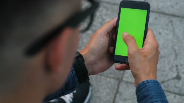 Close-up tiro de homem atraente nos óculos elegantes usando telefone celular com tela verde para conversar com amigos na cidade. Chave Chroma . — Vídeo de Stock