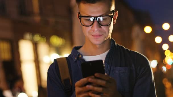 Hombre atractivo con gafas en la camisa de mezclilla usando un teléfono inteligente en la calle a la hora de la noche en la ciudad . — Vídeos de Stock