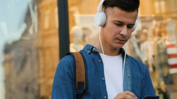 Hombre atractivo en las gafas con teléfono principal escuchando música en el fondo de la ciudad. Vista desde un lado. Dolly disparó . — Vídeo de stock