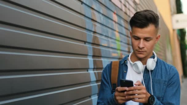Портрет привлекательного молодого человека с головным телефоном на шее, использующего мобильный телефон для общения с друзьями во время прогулки по улице. Вид сбоку . — стоковое видео