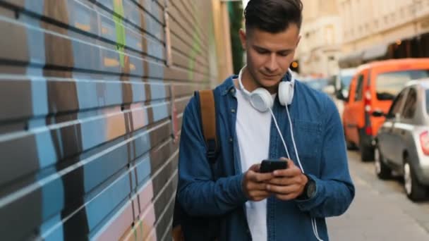Привлекательный молодой человек с головным телефоном на шее, использующий мобильный телефон для общения во время прогулки по улице . — стоковое видео