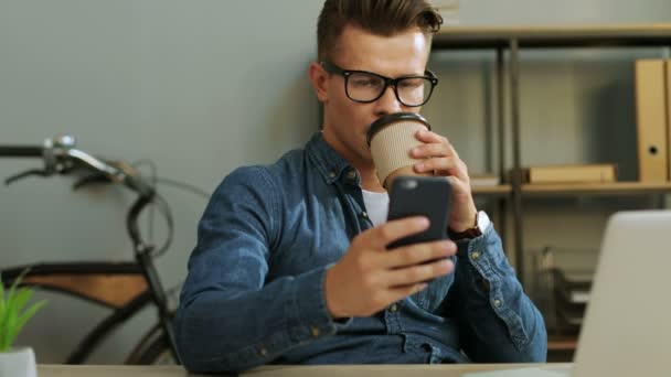 Jovem homem de negócios atraente usando telefone inteligente enquanto sentado no escritório e bebendo café . — Vídeo de Stock