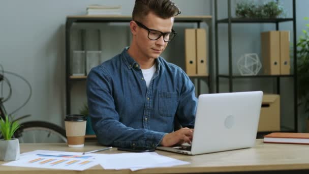 Привабливий молодий бізнесмен в окулярах працює в сучасному офісі, використовуючи ноутбук, дивлячись на камеру і посміхаючись . — стокове відео