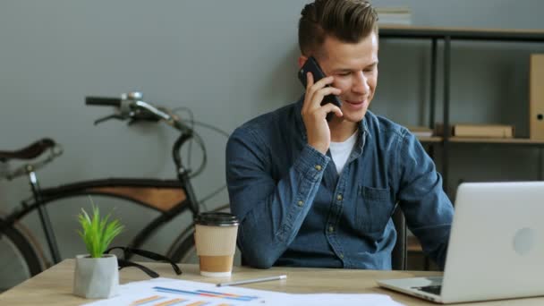 Porträt eines ernsthaften jungen Geschäftsmannes mit Brille, der im modernen Büro mit seinem Laptop arbeitet, während er mit dem Smartphone spricht. — Stockvideo