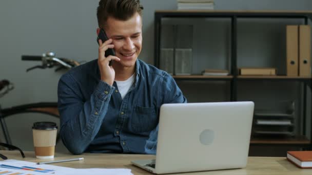 Atractivo joven hombre de negocios en las gafas que trabajan en la oficina moderna utilizando su computadora portátil mientras habla en el teléfono inteligente con el socio . — Vídeo de stock