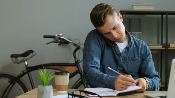 若いビジネス人スマート電話で話していると、事務所で作業しながらメモ帳に何かを書いて. — ストック動画