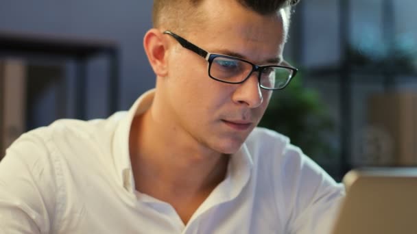 Primer plano retrato de un joven hombre de negocios con gafas en la camisa blanca trabajando en el portátil en la elegante oficina . — Vídeo de stock