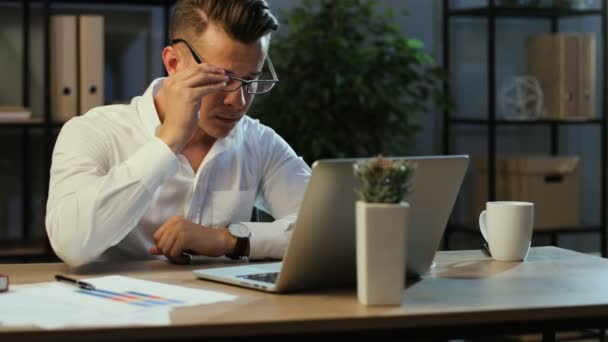 Attraktiv ung affärsman som bär glasögon och börja arbeta i moderna office använder laptop på kvällen. — Stockvideo