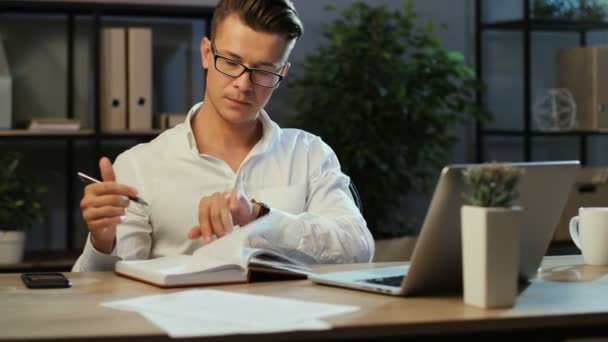 Joven hombre de negocios que usa el ordenador portátil para señalar algo en el cuaderno de notas mientras trabaja por la noche en la oficina . — Vídeos de Stock