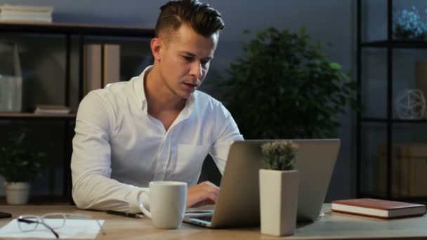 Aantrekkelijke jonge zakenman in klassieke shirt proberen werken met anoying hoofdpijn ter naar de laptop in het stijlvolle kantoor in de avond. — Stockvideo