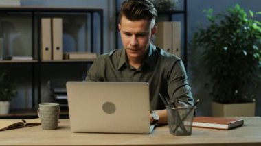 Ciddi genç iş adamı akşam anda modern ofis içinde belgili tanımlık laptop üzerinde çalışan ve çok yorgun hissediyorum haki gömlek.