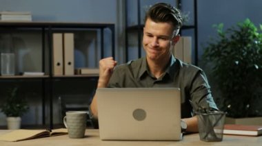 Onun iş ve resultats gurur bitirme mutlu genç iş adamı haki gömlek akşam anda modern ofis içinde belgili tanımlık laptop üzerinde çalışan.