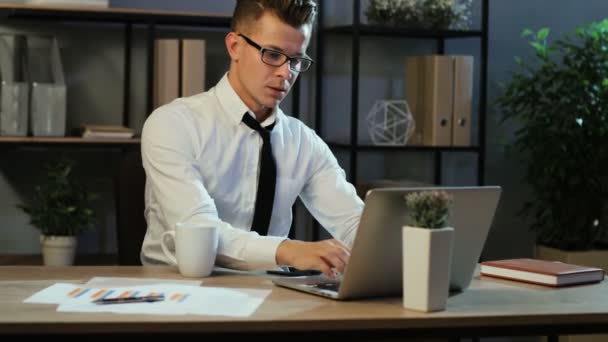 Homme d'affaires attrayant en chemise décontractée vérifiant le rapport financier et utilisant le téléphone intelligent dans le bureau le soir . — Video