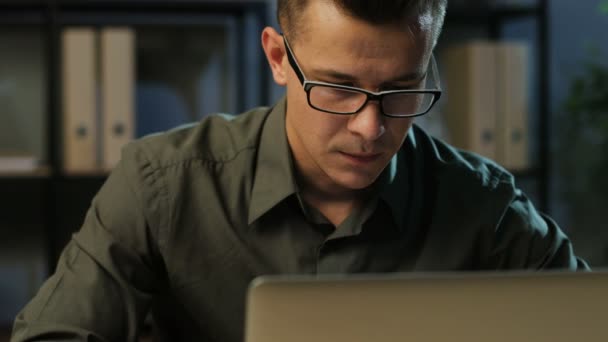 Retrato del joven hombre de negocios guapo con la camisa caqui trabajando en la computadora portátil en la oficina elegante y bostezando durante el tiempo de la noche . — Vídeos de Stock