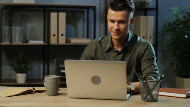 Glücklicher junger Geschäftsmann im khakifarbenen Hemd, der abends am Laptop im modernen Büro arbeitet. — Stockvideo