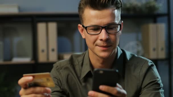 Retrato de hombre de negocios atractivo usando tarjeta de crédito y teléfono inteligente para compras en línea en el fondo de la oficina por la noche . — Vídeos de Stock