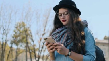 Bir smartphone aygıtında manifatura kadın. Dışında cep telefonu kısa mesaj yazarak şapka ve gözlük şehirde yürürken modern akıllı telefon kullanarak güzel genç kadın