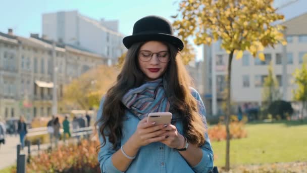 Close-up portret van lachende vrouw smartphone kijken naar de camera te houden. Jong meisje kijken camera bij herfst stad achtergrond. — Stockvideo