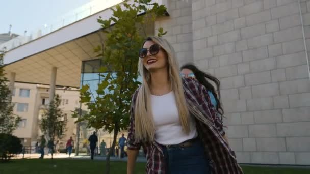 Dos jóvenes amigas felices dando a cuestas, riendo y caminando sobre el soleado fondo de la ciudad. Feliz joven novias abrazando — Vídeos de Stock