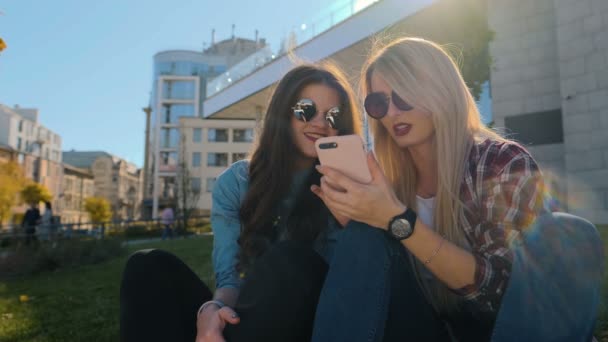 Retrato de dos chicas hipster atractivas en ropa casual relajarse y charlar. Mujer usando el teléfono inteligente escribiendo mensaje de texto fuera — Vídeos de Stock