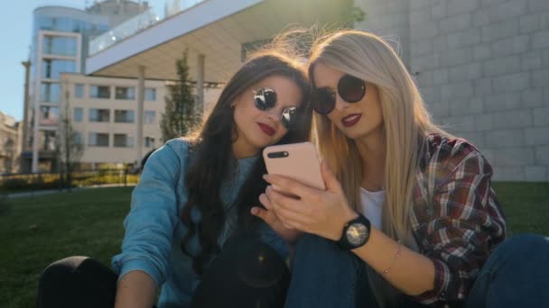 Retrato de duas meninas hipster felizes em roupas casuais relaxar e conversar mais. Mulher usando telefone inteligente digitando mensagem de texto fora. Eles estão felizes e sorrindo, sentados na grama — Vídeo de Stock