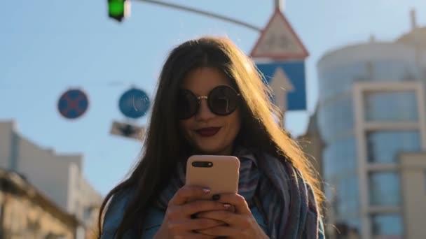 Primer plano retrato de una hermosa mujer joven caminando con un teléfono inteligente y mensajero de mensajes de texto. Ciudad de otoño en el telón de fondo — Vídeo de stock