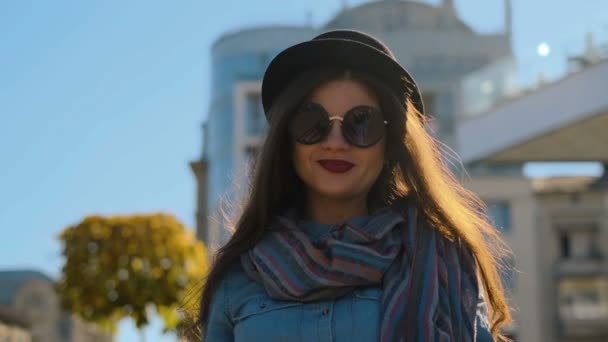 Closeup Πορτρέτο γυναίκας χαμογελαστά ντυμένοι μαύρο καπέλο και ήλιο γυαλιά περπάτημα και ψάχνει για την κάμερα. Νεαρό κορίτσι βλέπουν φωτογραφική μηχανή στο Φθινοπωρινό φόντο πόλη. — Αρχείο Βίντεο