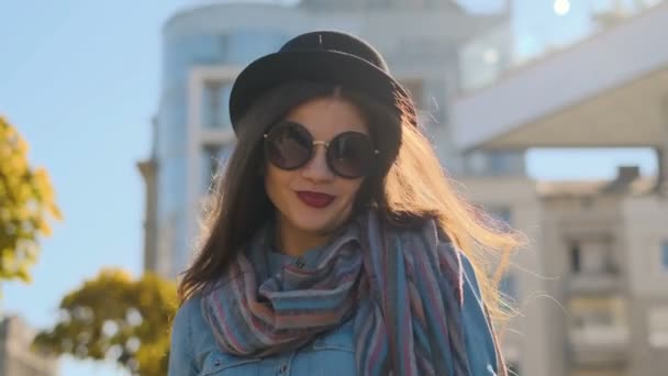 Retrato de cerca de una mujer sonriente vestida con sombrero negro y gafas de sol caminando y mirando a la cámara. Chica joven mirando a la cámara en el fondo de la ciudad de otoño . — Vídeo de stock