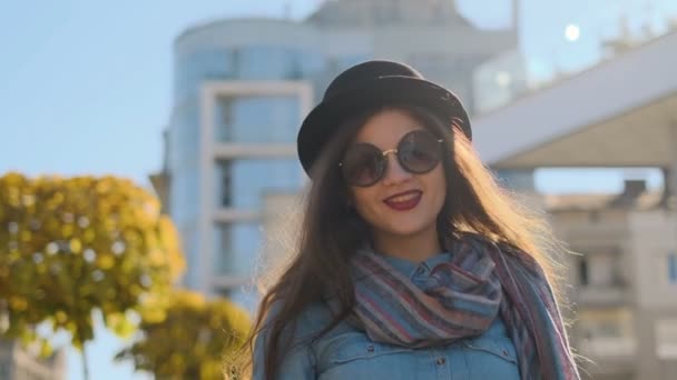 Retrato de mujer sonriente vestida con sombrero negro y gafas de sol caminando y mirando a la cámara. Chica joven mirando a la cámara en el fondo de la ciudad de otoño . — Vídeo de stock