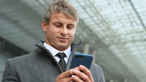 Zblízka portrétní sebevědomý mladý muž. Podnikatel v obleku drží chytrý telefon, SMS kolega a podíváme se na to přitom stát v lehké a moderní kancelářská budova — Stock video